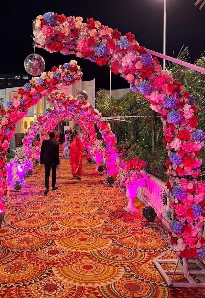 Tunnel of arches packed with pink, red and lilac flowers over a mandala-patterned walkway at night, with mirror balls hanging and guests walking through.