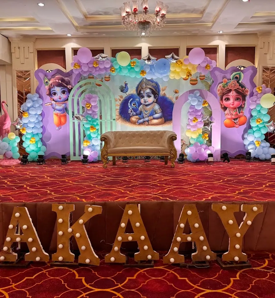Birthday stage inside the hall with a pastel balloon garland, silver star balloons, a gold settee and marquee-bulb letters glowing on the floor.