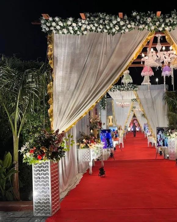Night entrance corridor of ivory curtains gathered on gold-trimmed peaked frames, with chandeliers and pastel lamps hanging above a red carpet.