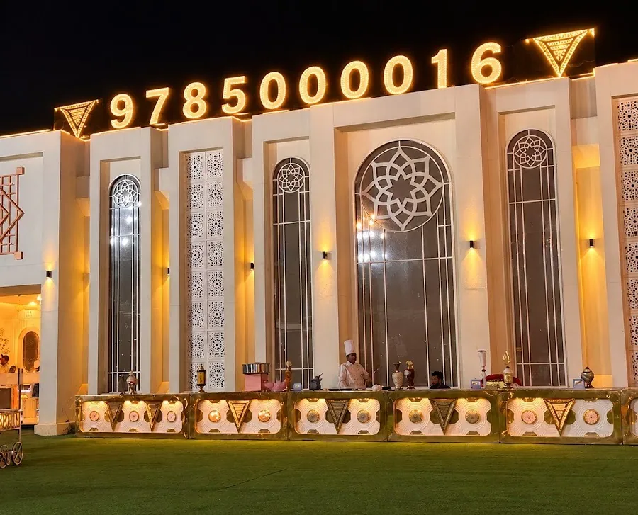 Madhavam's floodlit ivory facade at night: tall arched windows with star tracery, perforated jaali panels, and a marquee-bulb sign above the buffet counters on the lawn.