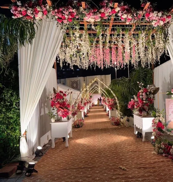 Night walkway under a canopy of pink and white flower strands, ivory drapes on both sides, leading to a tunnel of lit arches.