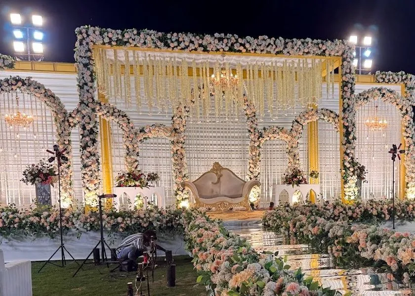 White and yellow floral wedding stage at night with arched floral frames, three chandeliers and a gold and cream settee behind mirror-topped tables.