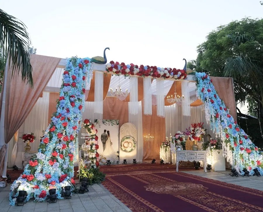 Outdoor reception stage at dusk with two cascading columns of blue hydrangea and red roses arching over a peach draped backdrop hung with chandeliers.