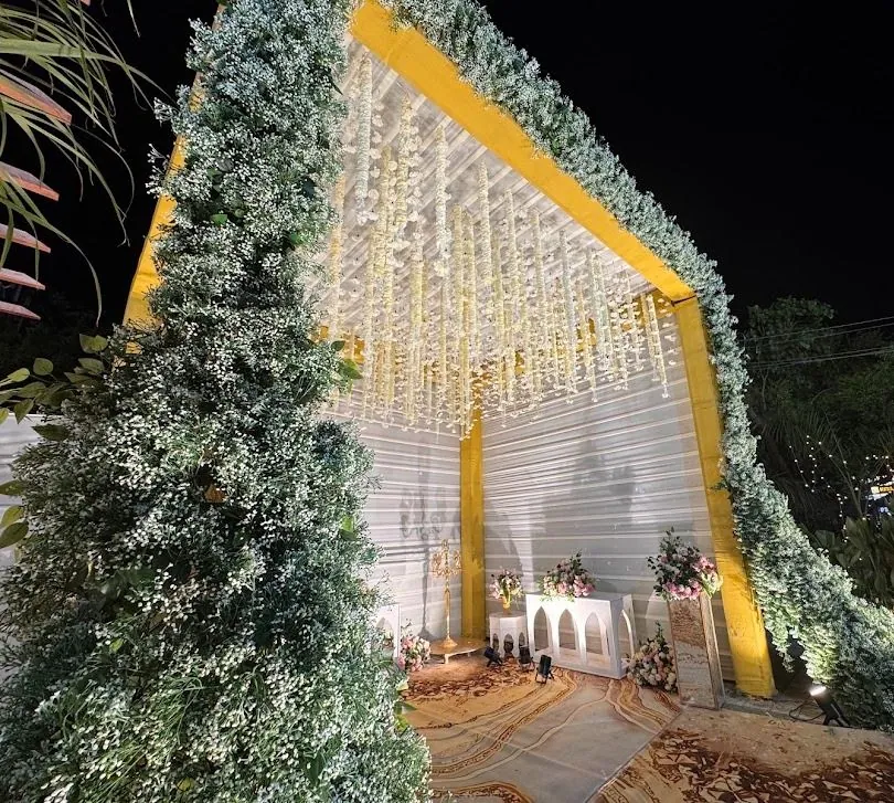 Night-lit A-frame welcome gate covered in white gypsophila and foliage, with strands of white and yellow flowers hanging from its ceiling.