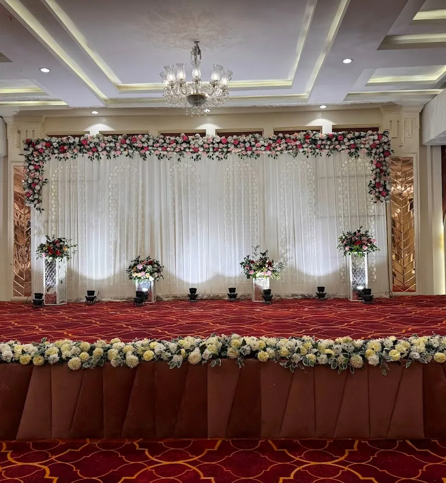 Hall stage with a long white sheer curtain wall topped by a rose garland, flanked by copper geometric panels, with floral pedestals and a crystal chandelier.