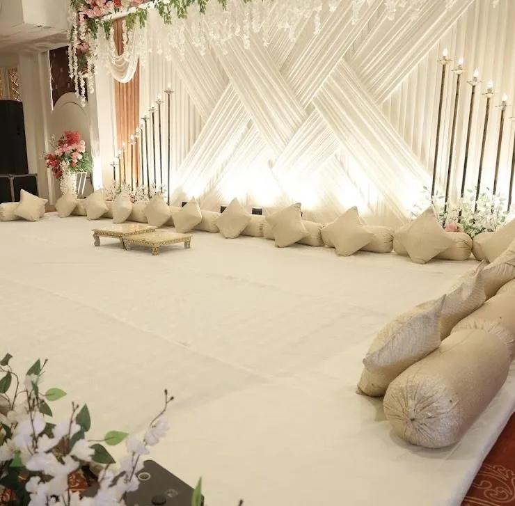 Indoor ceremony platform with cream bolsters and floor cushions, gold low tables, a backlit pleated backdrop and tall candelabra.
