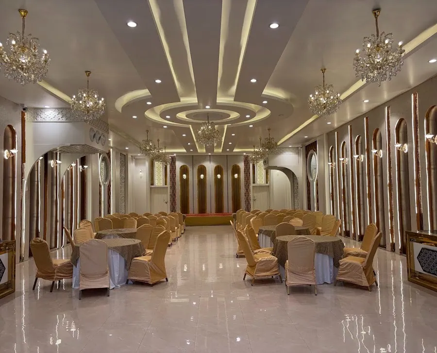 The main banquet hall at Madhavam: two rows of crystal chandeliers over polished cream marble, gold-framed arched mirror panels along both walls, round tables dressed in beige and gold leading to the stage.