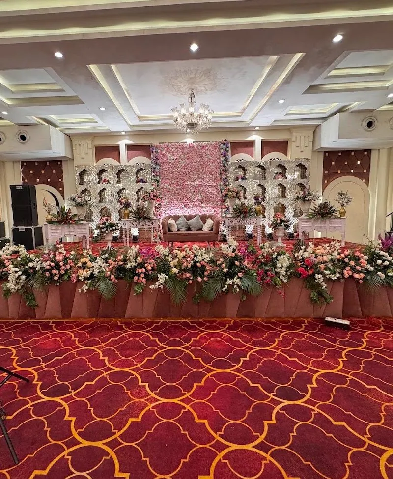 Hall reception stage with a pink and white floral wall flanked by white jaali arch screens, a blush loveseat and a crystal chandelier under the coffered ceiling.