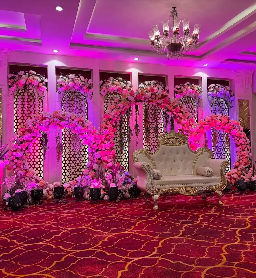 Dramatic magenta-lit stage: an S-curve of pink and white roses draped across tall white jaali arch screens, a cream and gold loveseat at centre, chandelier above.