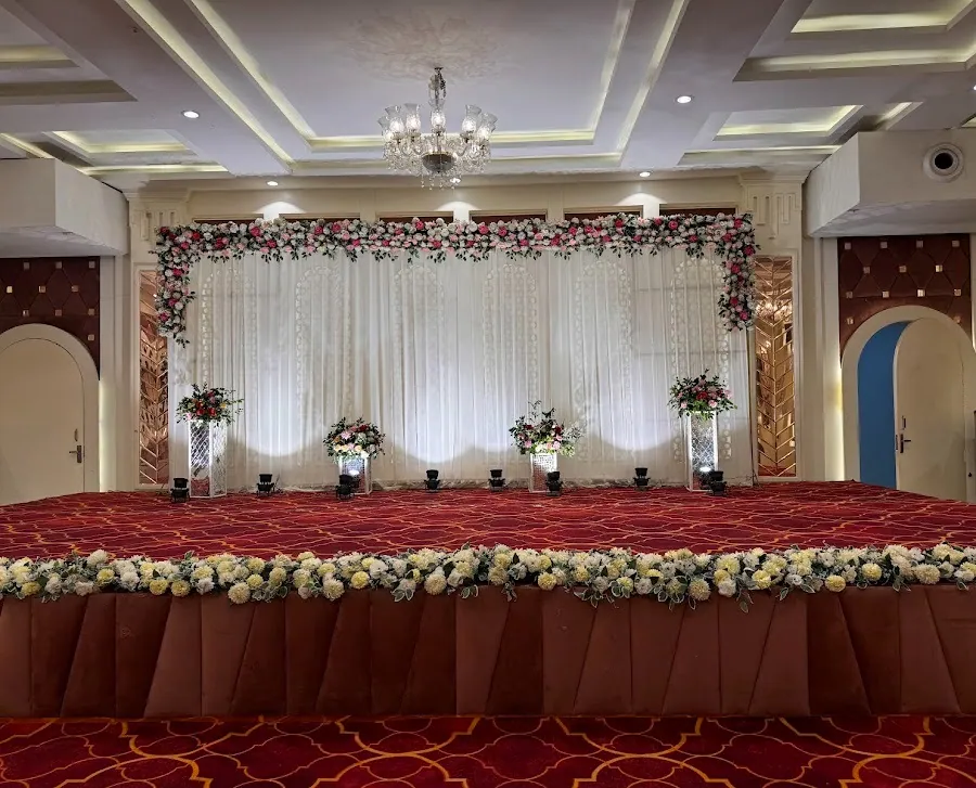 Indoor stage in the banquet hall with a cream curtain backdrop trimmed in pink and white roses, a crystal chandelier above and a red patterned carpet.