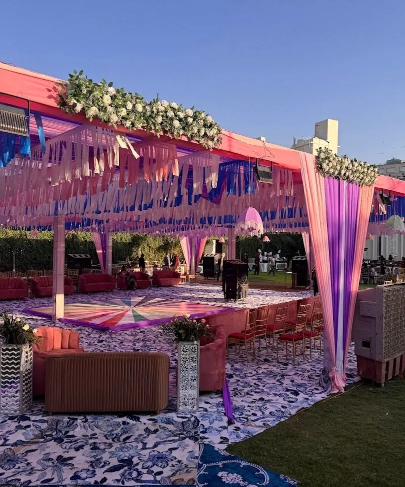 Open-sided lawn pavilion with coral and blue ribbon-fringe ceiling, gradient drape columns, velvet lounges, a starburst dance floor and rows of gold chairs.