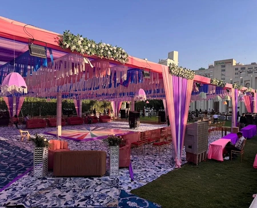 Daytime sangeet setup under a pink and purple draped canopy with hanging lampshades, a sunburst dance floor and lounge seating on the lawn.