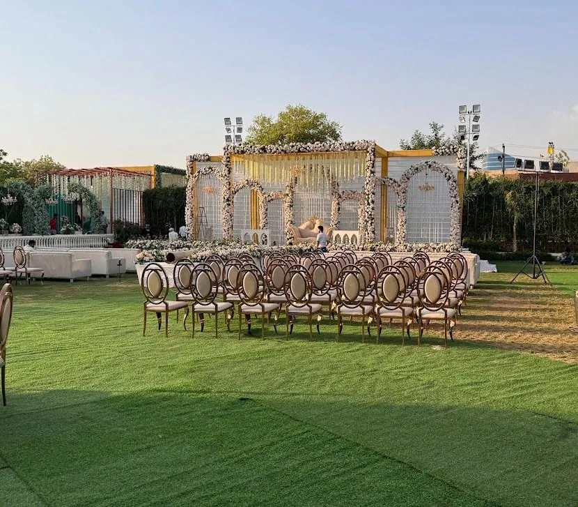 Daytime lawn wedding: white floral arch stage with gold trim and rows of gold chairs on the grass under a clear sky.