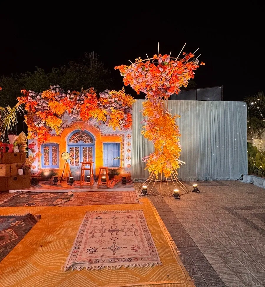 Mehndi photo set on the paved courtyard at night: a hand-painted haveli facade with a blue arched door, draped in orange foliage beside a bamboo teepee over layered kilim rugs.