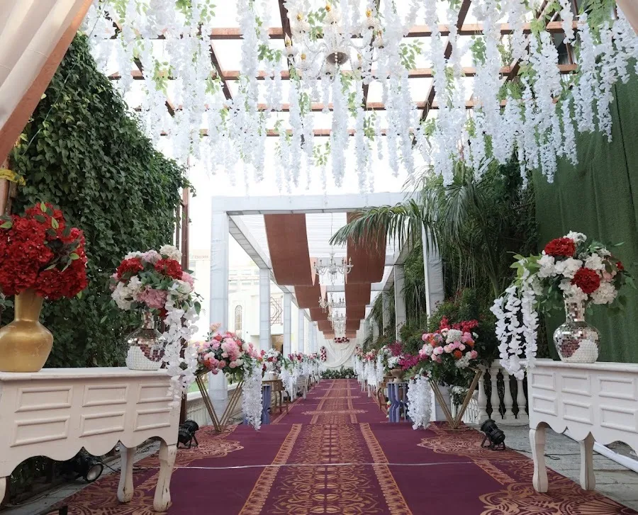Covered pergola walkway hung with trailing white wisteria and chandeliers, flanked by red and pink floral arrangements on white console tables, over a maroon and gold carpet.