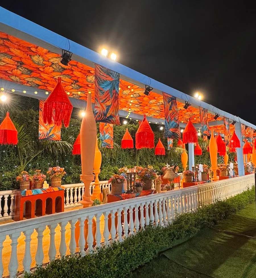 Covered terrace at night with a tropical-print canopy, red tasseled lanterns in a row, mustard vases along a white balustrade and string lights above.